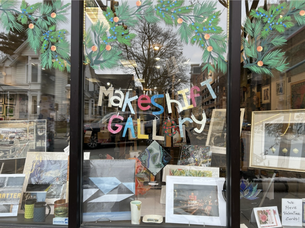 Image of Makeshift Gallery's front window, with the hand-made paper Makeshift Gallery sign in multiple colors hung above the arts display.