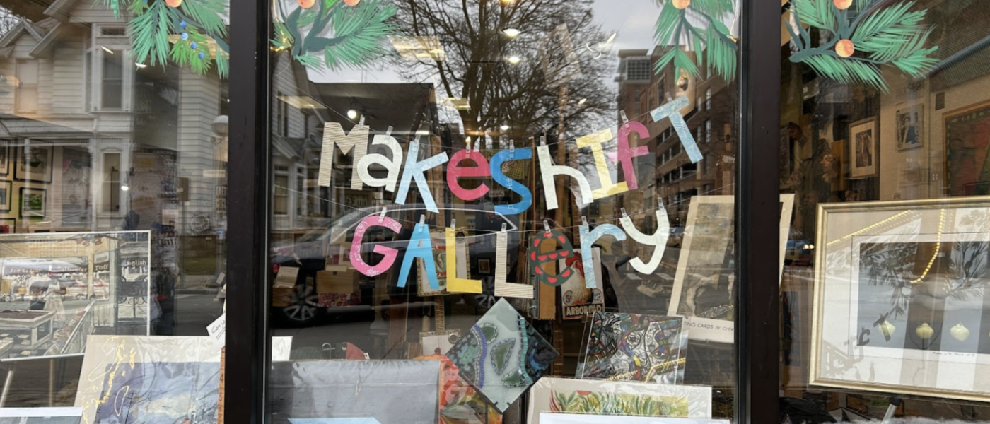 Image of Makeshift Gallery's front window, with the hand-made paper Makeshift Gallery sign in multiple colors hung above the arts display.